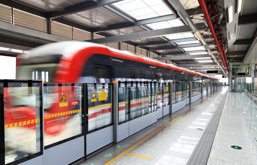 ​A motion-blurred red and black Beijing Subway train pulling into a modern station. The platform is equipped with grey and glass automatic platform screen doors featuring yellow warning stickers. The station has a high industrial ceiling with skylights and polished grey stone flooring with green directional arrows.