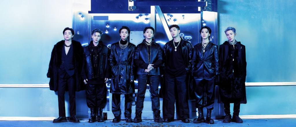 BTS members standing against a metallic vault door background, wearing sleek black leather and fur outfits.