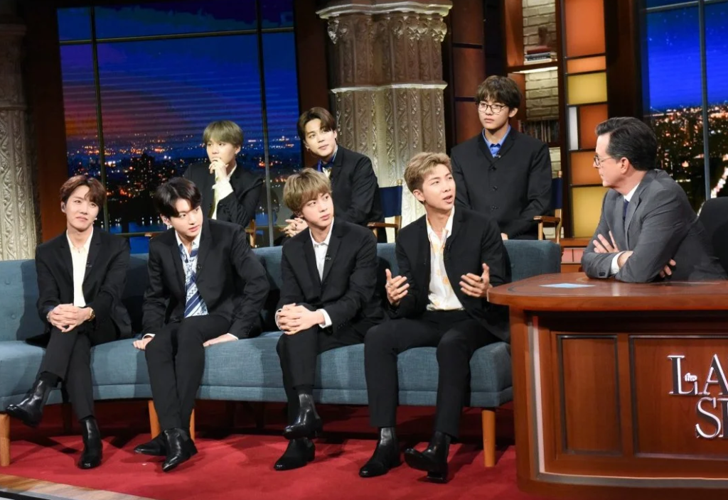 BTS members sitting on a sofa during an interview with Stephen Colbert in sleek black suits