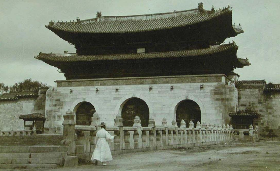 A historical photograph of Gwanghwamun Gate in the 1920s, featuring a person in traditional Hanbok walking in front of the stone Woldae and the wooden pavilion.