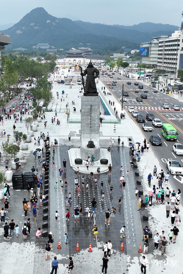 A high-angle view of Gwanghwamun Square featuring the statue of Admiral Yi Sun-sin and citizens enjoying the fountain on a summer day.