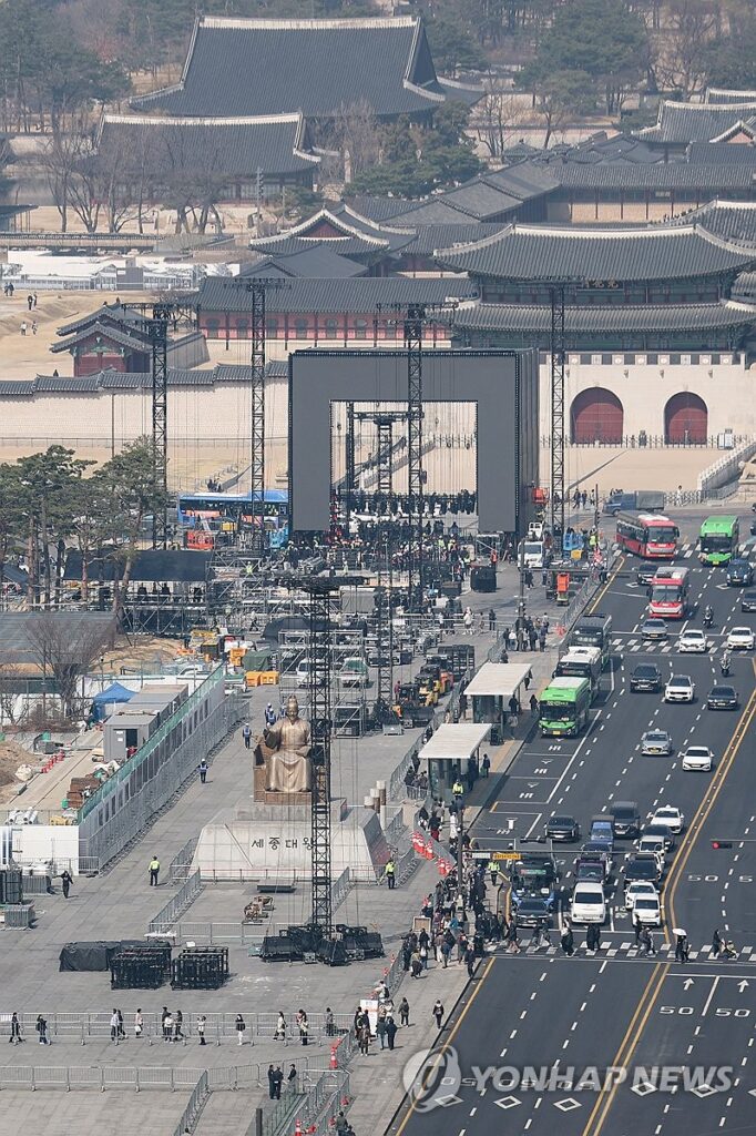 An aerial view of Gwanghwamun Square showing the massive stage construction for a BTS performance, featuring the King Sejong statue and Gwanghwamun Gate in the background