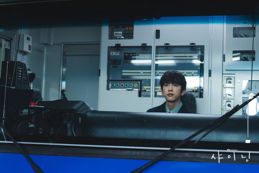 ​A cinematic close-up of actor Park Jin-young seated inside a Seoul Metro subway train cabin. He wears a navy uniform and light shirt, looking intently ahead. Behind him is a wall of complex electrical panels with buttons and indicator lights. A blue bus or train front panel and wiper are in the foreground. Korean text logo in the bottom right corner. The lighting is cool and blue.
