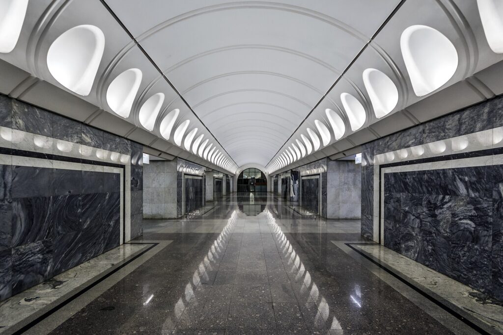 ​A wide-angle view of the grand interior of Komsomolskaya Station in the Moscow Metro. The yellow arched ceiling is decorated with elaborate white stuccos, mosaic panels, and massive bronze chandeliers. Marble pillars line the platform, and the floor is made of polished granite. The station looks like a luxurious palace ballroom.