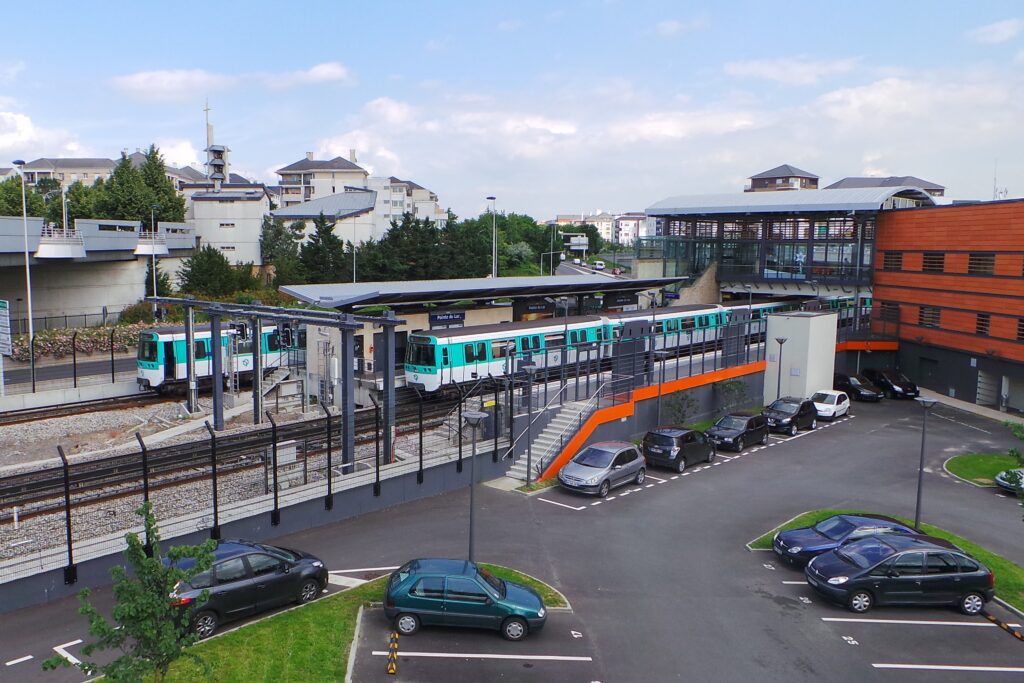 A mint-green and white Paris Métro train at an outdoor elevated station with a grey metal roof. A parking lot with several cars is visible in the foreground.