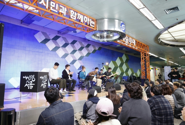 ​A dynamic photograph capturing a live musical performance on a professional stage built inside a bustling Seoul Metro station, labeled 'Art Stage' and 'Culture Event' in Korean and English. The band consists of four members: a male vocalist, an acoustic guitarist, an electric guitarist, and a keyboardist, all playing with enthusiastic expressions. Behind them, a large screen displays abstract geometric patterns in vibrant blue and green. In the background, clear LED screens show the next train arrival and station information. Surrounding the stage is a diverse, diverse crowd of smiling commuters—young students, families, and seniors—all stopped to watch, applaud, and record the performance on their phones. The overall feel is one of shared joy and community in a modern, clean underground public space, demonstrating why Seoul's system is a global favourite.