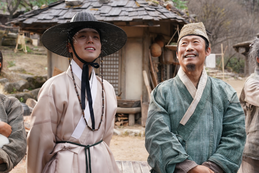 Two men in traditional Joseon Dynasty clothing (Hanbok) are standing outdoors and looking upward with bright smiles. The man on the left wears a light-colored robe and a black wide-brimmed hat (Gat), while the man on the right wears a rustic green robe and a simple headcloth.