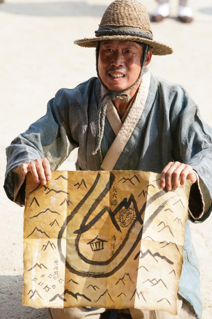 Actor Yoo Hae-jin, dressed in a rustic Joseon-era robe and a straw hat (Paerangi), is holding up a large, hand-drawn map. The map features traditional brush-painted mountains and a tiger illustration. He has an intense, slightly desperate facial expression.