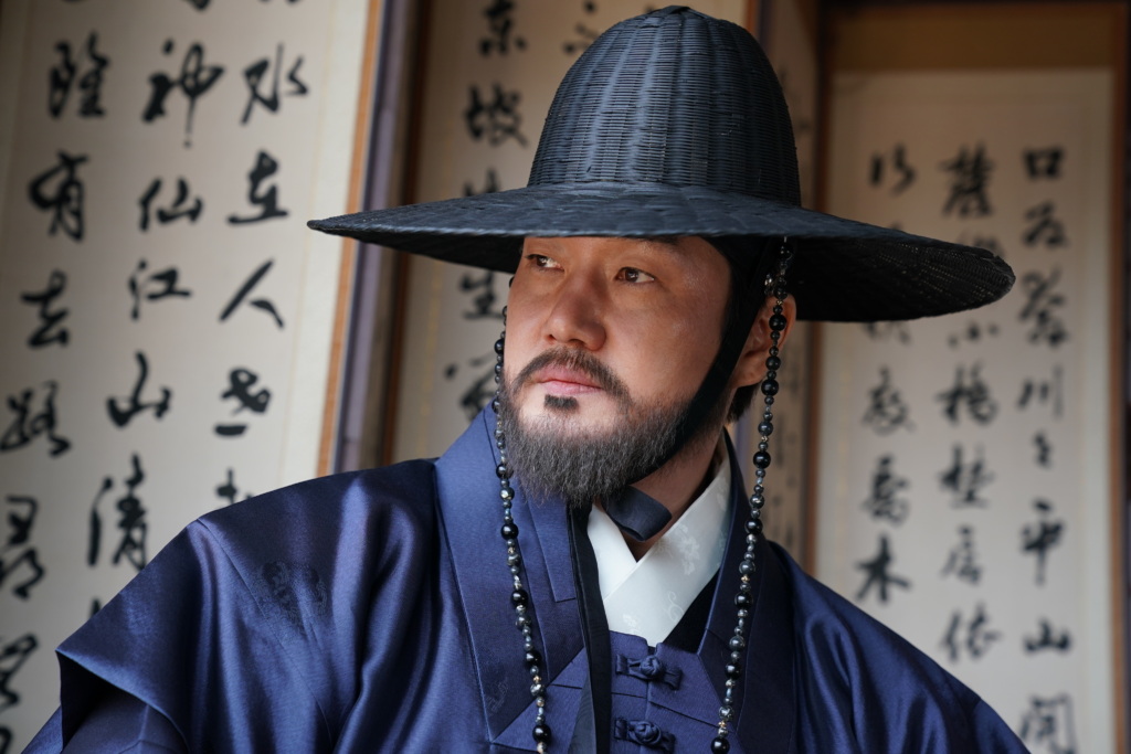 A close-up shot of actor Yoo Ji-tae in traditional Joseon Dynasty attire. He is wearing a dark navy silk robe (Hanbok) and a black wide-brimmed hat (Gat) with decorative bead strings. He has a well-groomed beard and a piercing, serious gaze. In the background, traditional Korean calligraphy screen is visible.