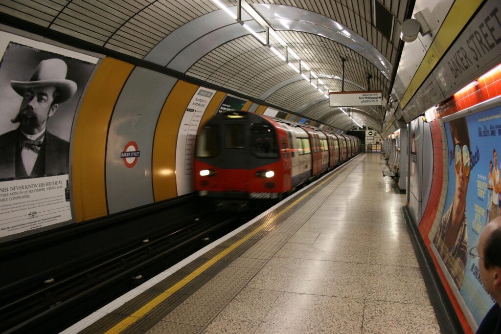 ​A motion-blurred red, white, and blue London Underground train entering a curved station platform at Baker Street. The platform features an arched tiled ceiling and large advertising posters on the walls. A sign indicates "Baker Street" and "Northbound platform 9." The overall atmosphere reflects the classic, deep-level tube station design.