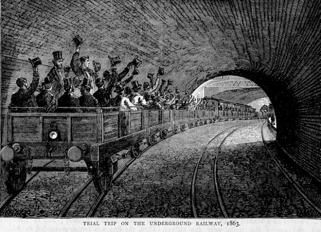 ​Black and white historical photograph of the first London Underground steam train at a station in 1863. A classic steam engine is on the tracks, surrounded by passengers in Victorian-era clothing on a gas-lit platform. The station has a high, arched brick ceiling with visible smoke from the locomotive.