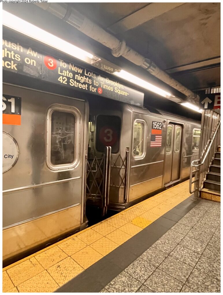 ​A silver stainless steel New York City Subway car (Line 3) at an underground station platform. A black overhead sign indicates the train's route to New Lots Av and Times Square. The platform has yellow tactile paving, and a staircase is visible on the right. The lighting is warm and slightly dim, typical of older NYC stations.
