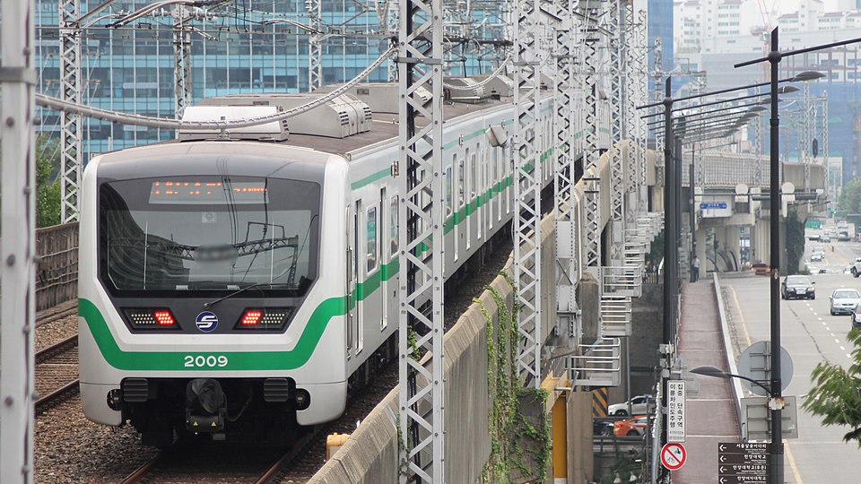 A sleek, modern Seoul Metro Line 2 train with a large, curved black glass front and bright LED headlights. The train's body is silver with a vibrant green stripe. It is stopped at a clean, well-lit underground station with glass screen doors. The overall look is futuristic and high-tech.