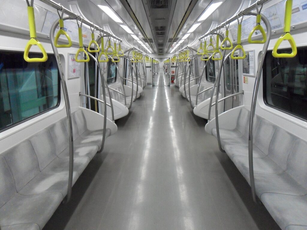 ​The wide and clean interior of a Seoul Metro Line 9 train. The cabin features beige walls, stainless steel grab bars, and dark grey seating. Digital information displays are mounted on the ceiling, and the floor is spotless. The lighting is bright and even, creating a comfortable atmosphere for passengers.