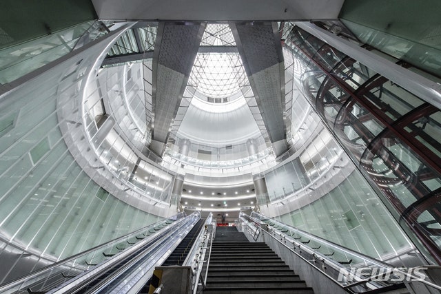 ​A dramatic, low-angle perspective looking upwards inside the massive, circular vaulted dome structure of Noksapyeong Station. Multiple overlapping concrete arches and metal beams create intricate geometric patterns, interspersed with large glass panels that allow natural sunlight to pour into the multiple circular, ramped levels far below. Below the tracks and platform levels, which are clean and active with Lines 6 trains, exhibit spaces for local artwork and sculptures are integrated into the station design. The overall feel is that of a modern underground cathedral, blending advanced transit technology with a serene, museum-like aesthetic.