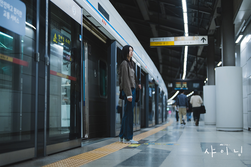 ​A cinematic high-quality still from the Korean drama 'Still Shining'. The female lead, a young woman with long dark hair wearing a casual jacket and jeans, stands alone on a clean and modern Seoul subway platform. She is looking slightly away with a contemplative and calm expression. Behind her is a sleek white and blue train (Line 4) with open screen doors. The platform is brightly lit with modern LED fixtures, and the floor is polished. In the background, a few blurred figures of other commuters can be seen, emphasizing the mix of solitude and public space. The drama title 'Still Shining' is written in elegant Korean script in the bottom right corner. The overall atmosphere is quiet, reflective, and beautifully composed.