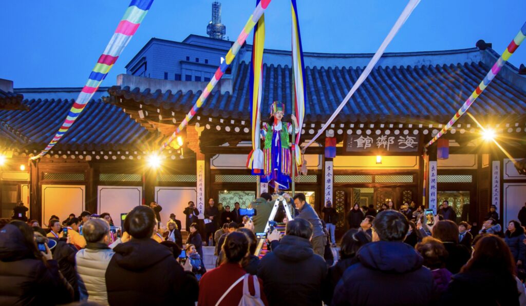 A vibrant evening scene of a traditional Korean shamanic ritual called 'Kkotmaji-gut,' featuring a shaman performing in front of a large crowd at a traditional Hanok setting, provided by the Korea Gut Research Association.