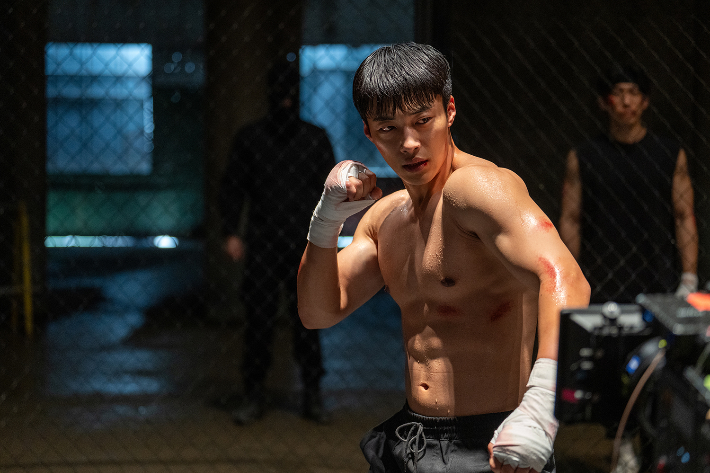 ​Actor Woo Do-hwan showing a muscular physique and boxing stance in a gritty arena for Netflix series Bloodhounds 2.